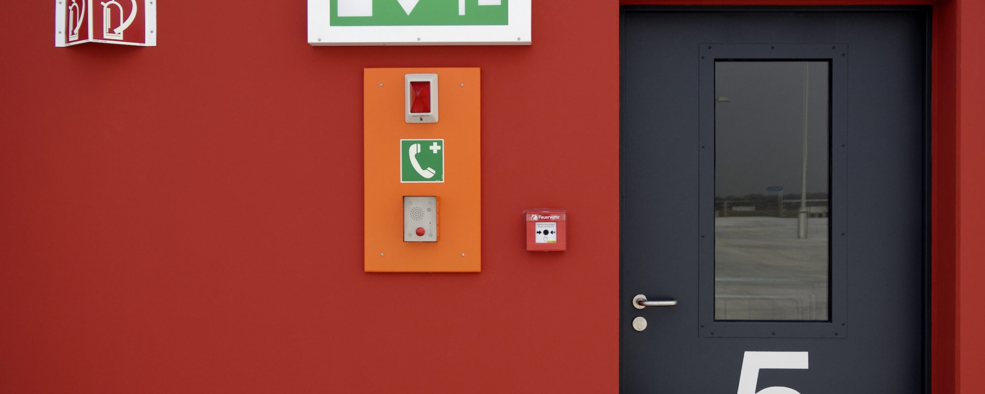 Green emergency exit sign, sos intercom, fire alarm button, pictogram for a fire extinguisher and ablack door with number 5 on a red wall.